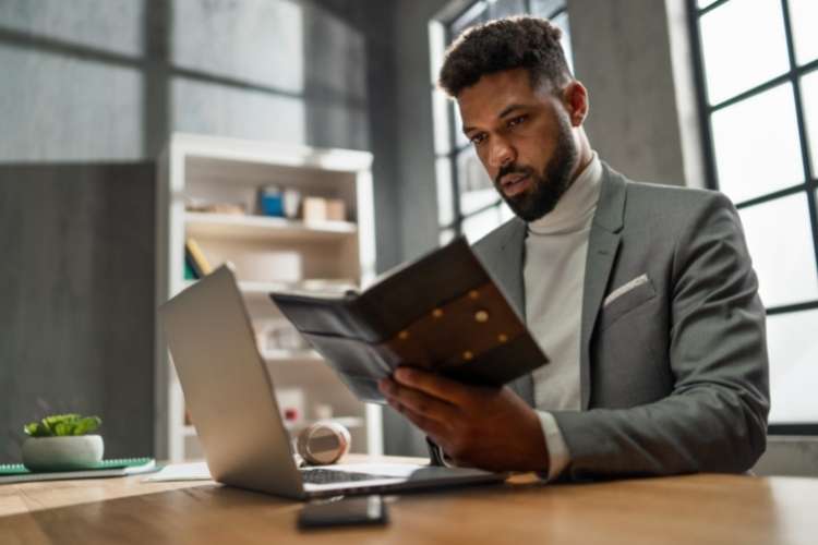 Homem negro de terno lendo anotações de caderno, com notebook à sua frente.