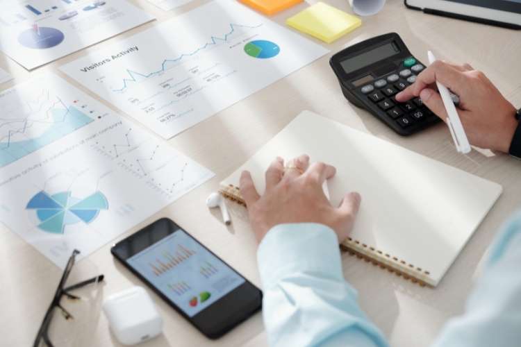 Pessoa analisando dados em papéis, fazendo contas e anotando em caderno.