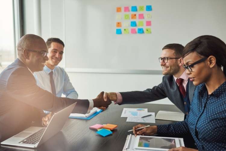 Quatro pessoas sentadas à mesa com itens de escritório à frente. Duas delas estão apertando as mãos, fechando negócio.